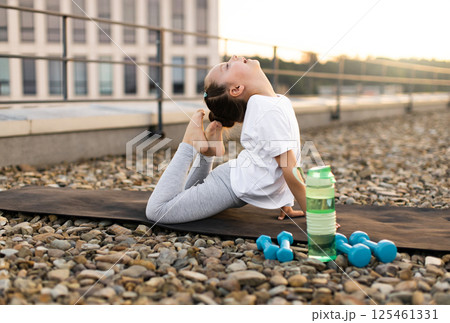 Young girl practicing yoga on rooftop during sunset, illustrating flexibility, balance, and fitness. Surroundings highlight urban outdoor environment with fitness essentials. 125461331