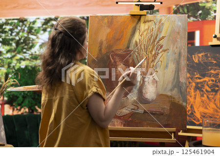 In a rustic workshop, artist mixes a variety of watercolor paint on a palette to create a colorful masterpiece on canvas. Woman painting with a brush and showing her skill, artwork creation. 125461904