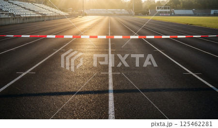 Running track with finish line barrier, empty stadium seating on the left, green grass and scoreboard on the right, bathed in warm sunlight, lines and lanes visible 125462281