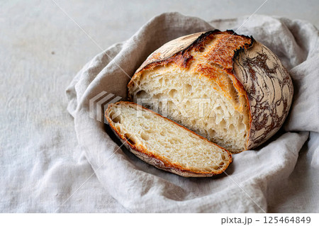 A sliced sourdough loaf with a crunchy crust reveals its airy texture, resting on a linen cloth, fresh from the oven 125464849