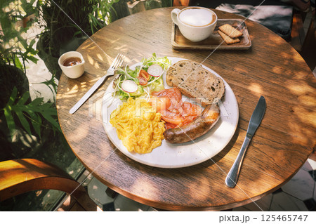 Scrambled eggs, fried pork sausage and sliced smoked salmon slices with rye bread toast and salad with radishes and Coffee with biscuits. A fresh breakfast in the sunshine outside. 125465747