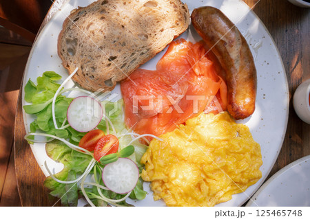 Breakfast platter featuring smoked salmon, scrambled eggs, sausage, vegetables, and whole grain bread served on a sunny morning at a local cafe close-up, top view Breakfast platter featuring smoked salmon, scrambled eggs, sausage, vegetables, and whole grain bread served on a sunny morning at a local cafe close-up, top view 125465748