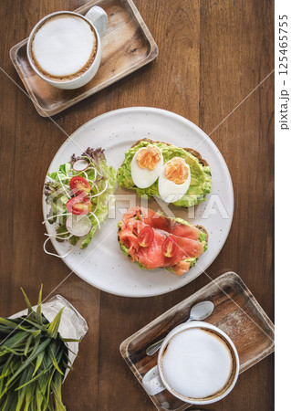 Toasts with avocado, smoked salmon and boiled eggs, salad and 2 cups of cappuccino coffee on a wooden table top view flatlay. Healthy and delicious breakfast for two with coffee or coffee break. 125465755