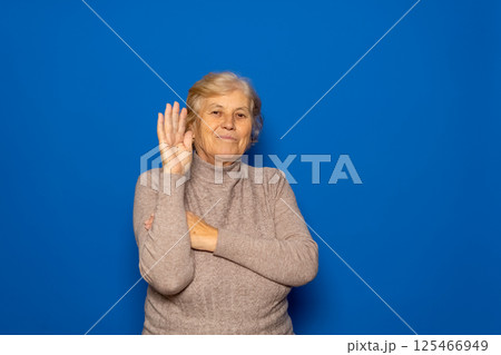 Hello concept. A happy, smiling, elderly Caucasian woman in her 70s waves with her hand outstretched. Isolated on a blue studio background. 125466949