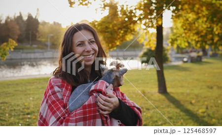 A joyful woman, cozy in a blanket, smiles while holding her pet in a serene autumn park with vibrant colors A joyful woman, cozy in a blanket, smiles while holding her pet in a serene autumn park with vibrant colors 125468597