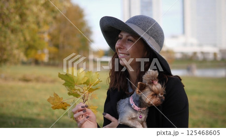A woman stylishly dressed in a hat is holding colorful leaves while cuddling her small dog in a serene park setting A woman stylishly dressed in a hat is holding colorful leaves while cuddling her small dog in a serene park setting 125468605