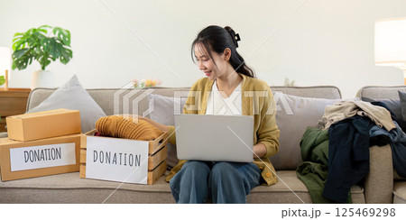 Woman sitting on the couch with a laptop and donation boxes, organizing clothes and smiling in a cozy living room Woman sitting on the couch with a laptop and donation boxes, organizing clothes and smiling in a cozy living room 125469298