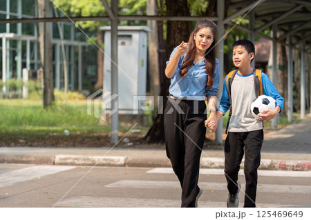 Playful Anticipation on the Way to School. A mother and her eager son walking hand in hand, soccer ball in hand. 125469649