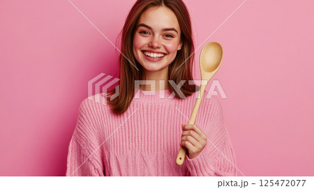 Cheerful young woman holding a wooden spoon against a pink background Cheerful young woman holding a wooden spoon against a pink background 125472077