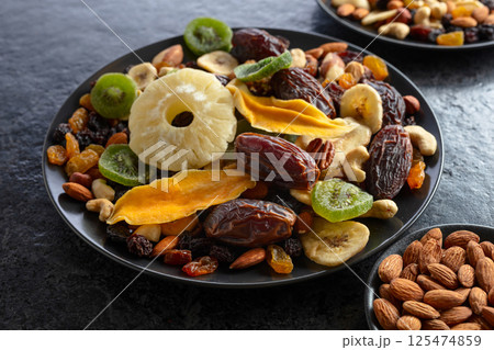 Dried tropical fruits, nuts, and raisins in black plate. 125474859