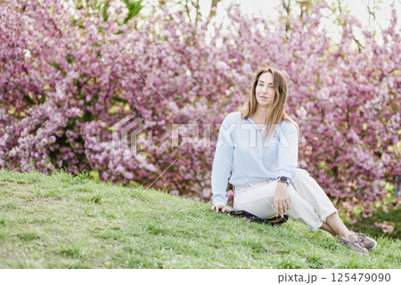 Smiling woman standing among blooming pink cherry blossom sakura trees wearing a light blue shirt and enjoying a bright spring day outdoors 125479090