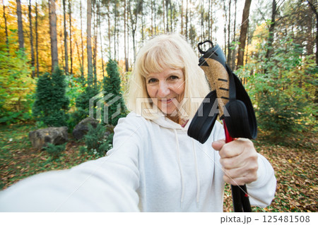 Senior woman taking a cheerful selfie during a nordic walk and take selfie on smartphone. Concept of active lifestyle, adventure, fitness, freedom and joy in the golden autumn forest 125481508