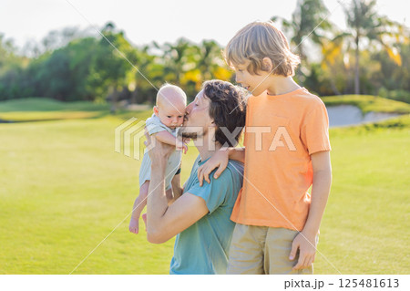 Father, older son, and newborn baby on a meadow in the park. Family spending quality time together in nature. Parenthood, love, and family bond concept Father, older son, and newborn baby on a meadow in the park. Family spending quality time together in nature. Parenthood, love, and family bond concept 125481613