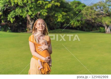 Mother holding her little baby in a yellow sling in the park. Warm and loving family moment. Babywearing, parenting, and mother-child bonding concept Mother holding her little baby in a yellow sling in the park. Warm and loving family moment. Babywearing, parenting, and mother-child bonding concept 125482909