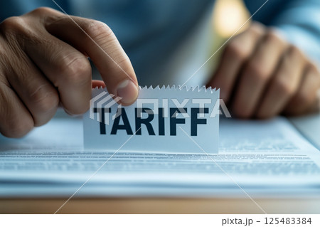 A close-up of a hand holding a paper labeled TARIFF above a document, symbolizing trade policies. The soft background suggests a serious yet focused mood, emphasizing economic discussions. 125483384