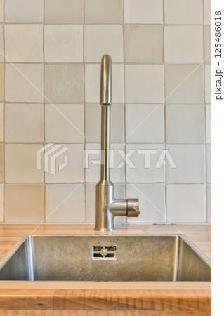 A sleek silver kitchen sink featuring a contemporary faucet against a light-colored tiled wall, showcasing minimalist design and functionality. 125486018