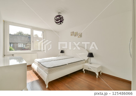 Bright and airy modern bedroom featuring a minimalist design with a white bed, wooden flooring, and a window allowing natural light to illuminate the space. 125486066