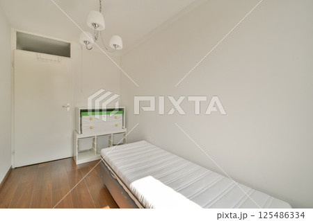 A bright and minimalist bedroom featuring a single mattress on a wooden floor, complemented by a modern chandelier and a compact storage unit. 125486334
