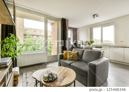 A bright and airy modern living room featuring a comfortable sofa, wooden coffee table, and a view of greenery through large windows, creating a welcoming atmosphere. A bright and airy modern living room featuring a comfortable sofa, wooden coffee table, and a view of greenery through large windows, creating a welcoming atmosphere. 125486346