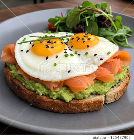A slice of avocado toast is topped with smoked salmon and two fried eggs, served with a small salad 125487905