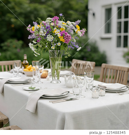 A table is set for an outdoor meal with a large vase of wildflowers in the center, near a white building 125488345