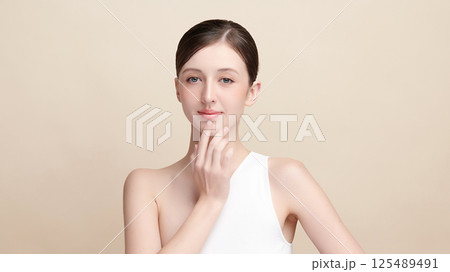 A beautiful young woman with clean, fresh skin against a beige background. Skincare, facial treatment, cosmetology, beauty, and spa. An elegant portrait of an woman. 125489491