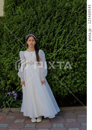 A young girl stands immersed in the bright moment before her First Communion. The light dress flows gently to the ground. A young girl stands immersed in the bright moment before her First Communion. The light dress flows gently to the ground. 125489545