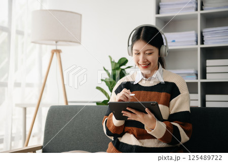 Smiling girl relaxing at home, she is playing music using smartphone tablet, and wearing white headphones. on sofa Smiling girl relaxing at home, she is playing music using smartphone tablet, and wearing white headphones. on sofa 125489722