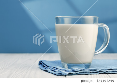 Glass mug brimming with fresh milk, resting on blue napkin atop white wooden table Glass mug brimming with fresh milk, resting on blue napkin atop white wooden table 125491249