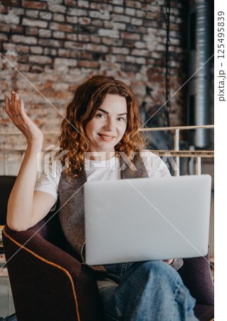 A young woman with curly red hair sits in a cafe with her laptop, looking confidently at the camera. Confident woman, modern entrepreneur, startup mindset, driven professional... 125495839