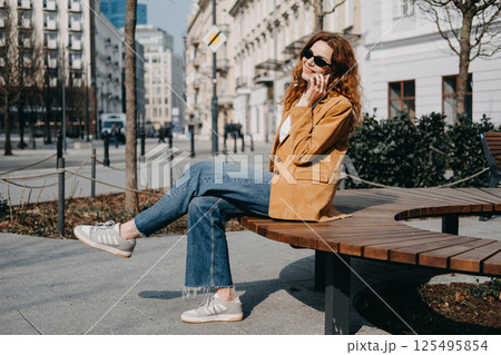 Young woman sitting on a curved wooden bench in the city while talking on her phone. Hybrid work, remote flexibility, digital workspaces, mobile professional... 125495854