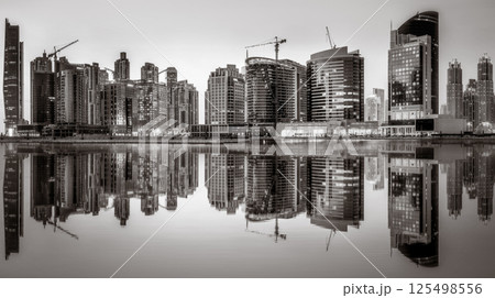 Monochrome city skyline with tall skyscrapers reflecting on calm water under a cloudy sky, Dubai Marina bay UAE 125498556