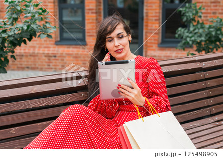 Woman sitting on bench with shopping bags and tablet. Digital lifestyle and modern shopping experience. Woman sitting on bench with shopping bags and tablet. Digital lifestyle and modern shopping experience. 125498905