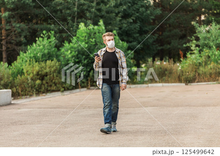 Man wearing a mask while walking in the park and checking his smartphone. Concept of modern life, health safety, and communication during pandemic times. Copy space 125499642