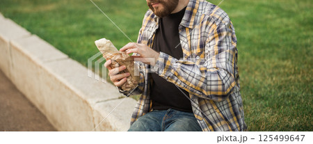 Man holding a street food wrap in craft paper outdoors. Concept of fast food, casual lifestyle, and outdoor snacking in urban park settings. Banner with copy space 125499647