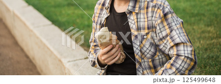 Man holding a street food wrap in craft paper outdoors. Concept of fast food, casual lifestyle, and outdoor snacking in urban park settings. Banner with copy space 125499648
