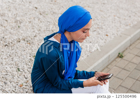 Young arab woman wearing hijab using smartphone while sitting outdoors. Technology, communication, and cultural identity concepts. 125500496