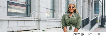 Confident african american woman standing against city architecture smiling. Concept of positivity, urban lifestyle and self-confidence. Banner with copy space 125501038