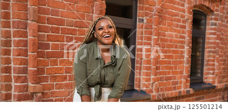 African american woman laughing joyfully while standing on a city street on a sunny day. Freedom, positivity, and happiness in an urban environment. Banner with copy space 125501214