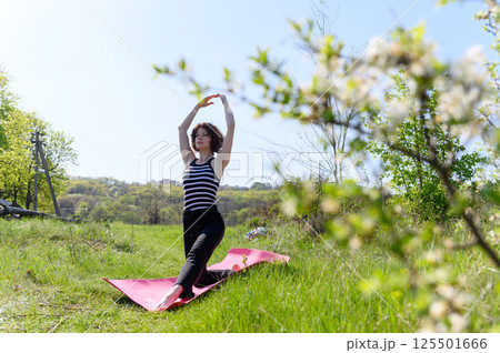 Woman practicing low lunge pose outdoors in spring garden with blooming trees. Woman practicing low lunge pose outdoors in spring garden with blooming trees. 125501666