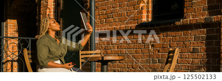 African american woman smiling and holding a tablet while sitting on outdoor cafe. Communication, happiness, and digital connection with loved ones. Banner with copy space 125502133