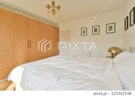 A stylish bedroom featuring a large bed, a wooden wardrobe, and decorative wall art, bathed in natural light for a serene atmosphere. A stylish bedroom featuring a large bed, a wooden wardrobe, and decorative wall art, bathed in natural light for a serene atmosphere. 125502596