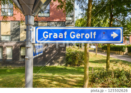 A blue directional sign indicating the location 'Graaf Gerolf' situated in an urban area with greenery and modern buildings in the background. A blue directional sign indicating the location 'Graaf Gerolf' situated in an urban area with greenery and modern buildings in the background. 125502626