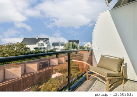 A beautifully designed modern balcony offers a comfortable chair and stunning views of a lush landscape and distant homes under a bright blue sky. 125502633