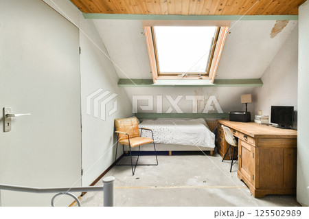 A simple yet elegant bedroom featuring a bed, desk, and chair, illuminated by natural light streaming through a skylight. The design is uncluttered and calming. 125502989