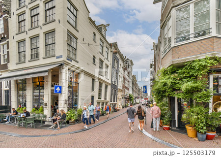 A picturesque street in Amsterdam featuring historic architecture, outdoor dining, and pedestrians enjoying the lively atmosphere on a sunny day. 125503179