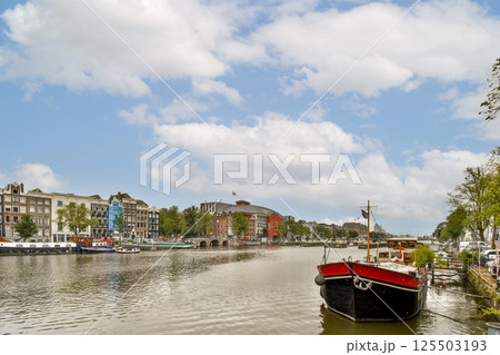 A picturesque view of an Amsterdam canal with charming buildings and a boat on the water. The serene atmosphere is enhanced by the blue sky and fluffy clouds. A picturesque view of an Amsterdam canal with charming buildings and a boat on the water. The serene atmosphere is enhanced by the blue sky and fluffy clouds. 125503193