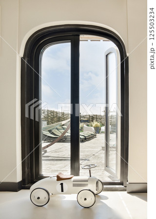 An elegant interior featuring a stylish toy car placed by a large window, overlooking a beautiful outdoor terrace and bright sky. An elegant interior featuring a stylish toy car placed by a large window, overlooking a beautiful outdoor terrace and bright sky. 125503234