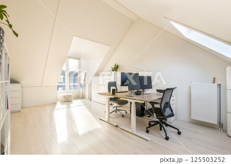 Bright and airy modern office space featuring sleek furniture, dual computer setups, and large windows allowing ample sunlight. A stylish, minimalistic design adds to productivity. 125503252