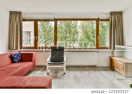 A contemporary living room featuring a red sofa, wooden furniture, and large windows overlooking greenery. The space is well-lit and inviting, blending comfort with style. 125503527